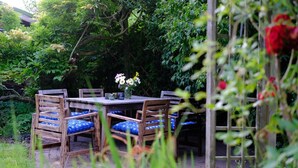 An outdoor wooden table with chairs, surrounded by lush greenery and a wooden trellis with red flowers.