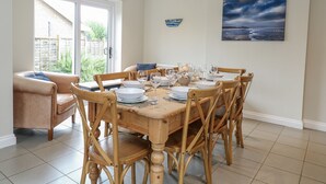 A dining area with a wooden table set for four, surrounded by wooden chairs. There is a view of a garden through a glass door, and a painting of a seascape on the wall. - Woodlark (King's Lynn)
