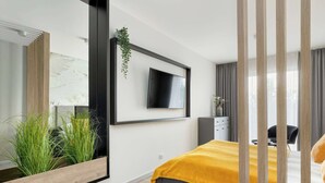 A modern hotel room with a large bed, a flat-screen TV mounted on the wall, a wooden bedside table, and a potted plant.