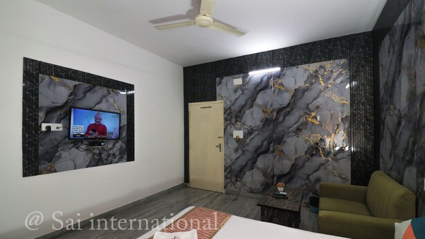 A modern hotel room with a flat-screen TV mounted on the wall, a ceiling fan, a bed with a neatly made bedspread, and a seating area with a sofa and armchair. - Sai International (Bidhannagar)