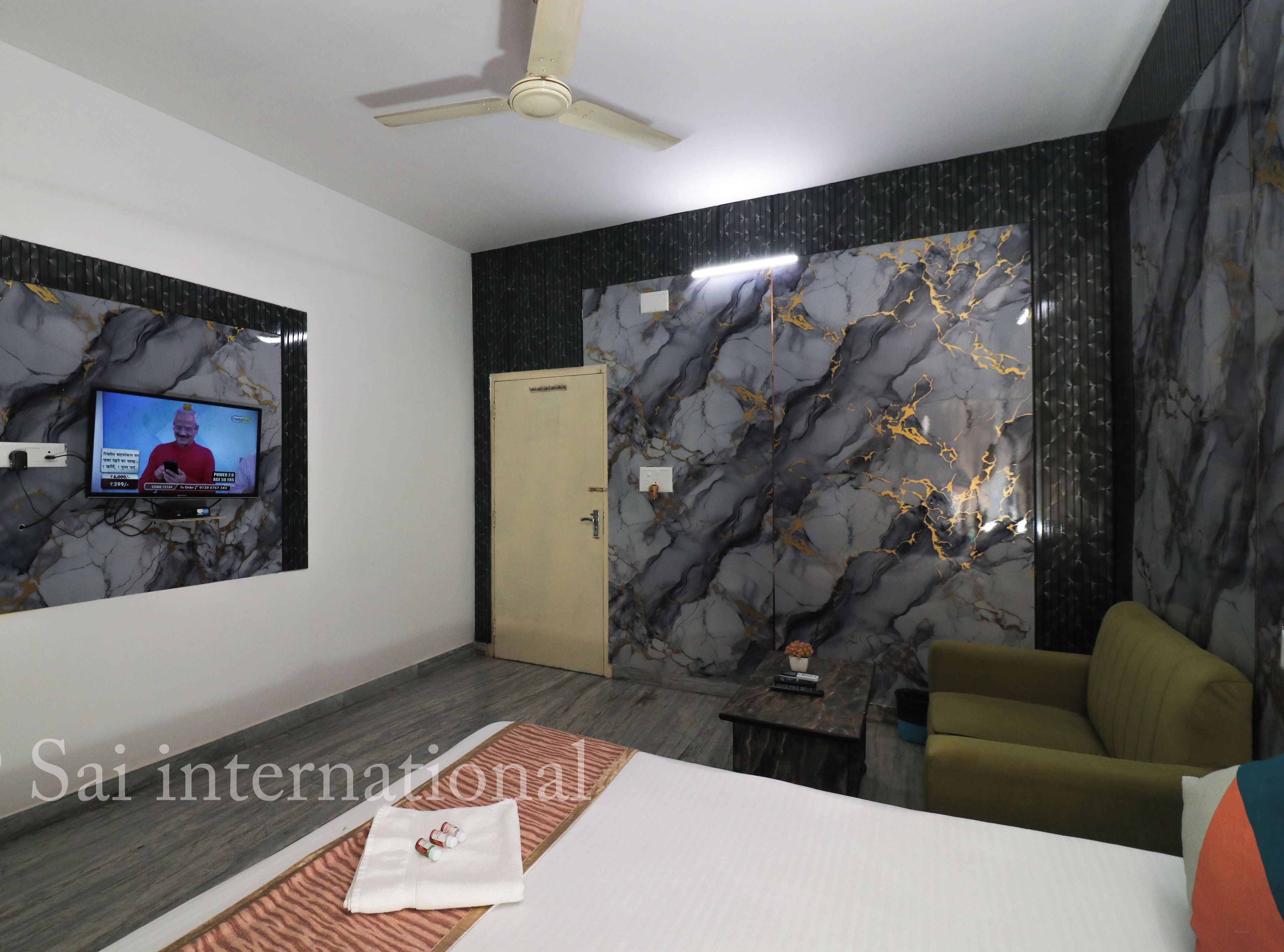 A modern hotel room with a flat-screen TV mounted on the wall, a ceiling fan, a bed with a neatly made bedspread, and a seating area with a sofa and armchair.
