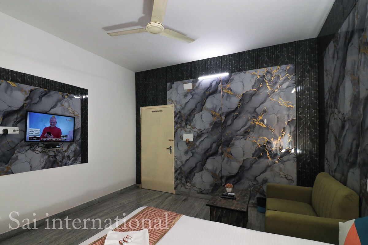 Una habitación de hotel moderna con un televisor de pantalla plana montado en la pared, un ventilador de techo, una cama con colcha impecable y una zona de estar con sofá y sillón.