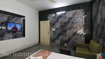 A modern hotel room with a flat-screen TV mounted on the wall, a ceiling fan, a bed with a neatly made bedspread, and a seating area with a sofa and armchair.