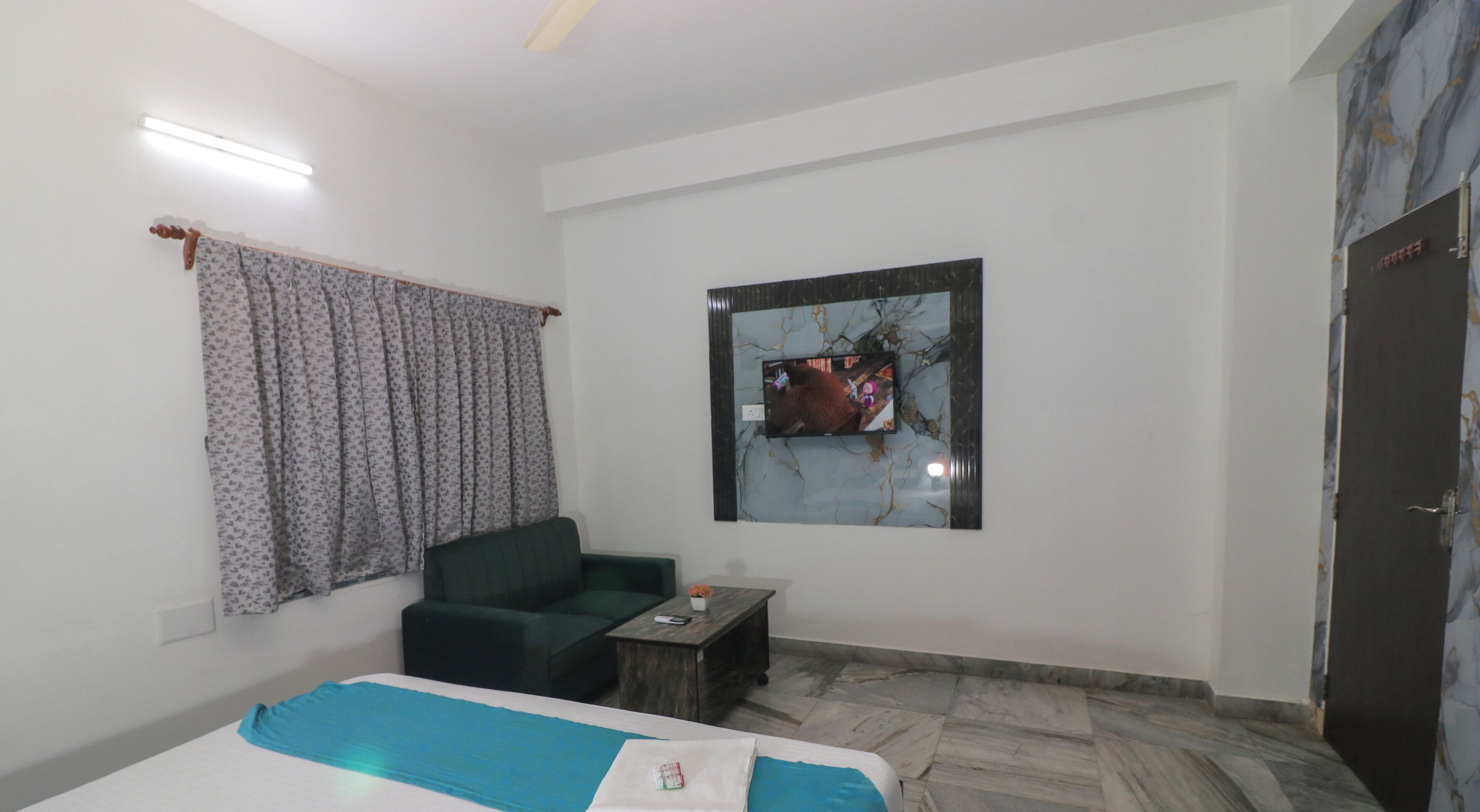 A hotel room with a bed, a green sofa, a small table, a TV mounted on the wall, and a door.