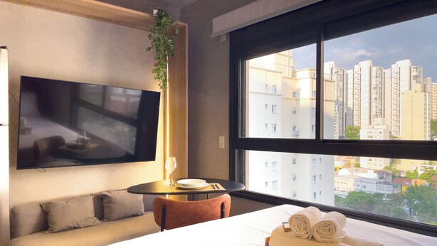 A hotel room with a bed, a TV mounted on the wall, a small table with a glass, and a view of high-rise buildings. - Lira Perdizes (Sao Paulo)