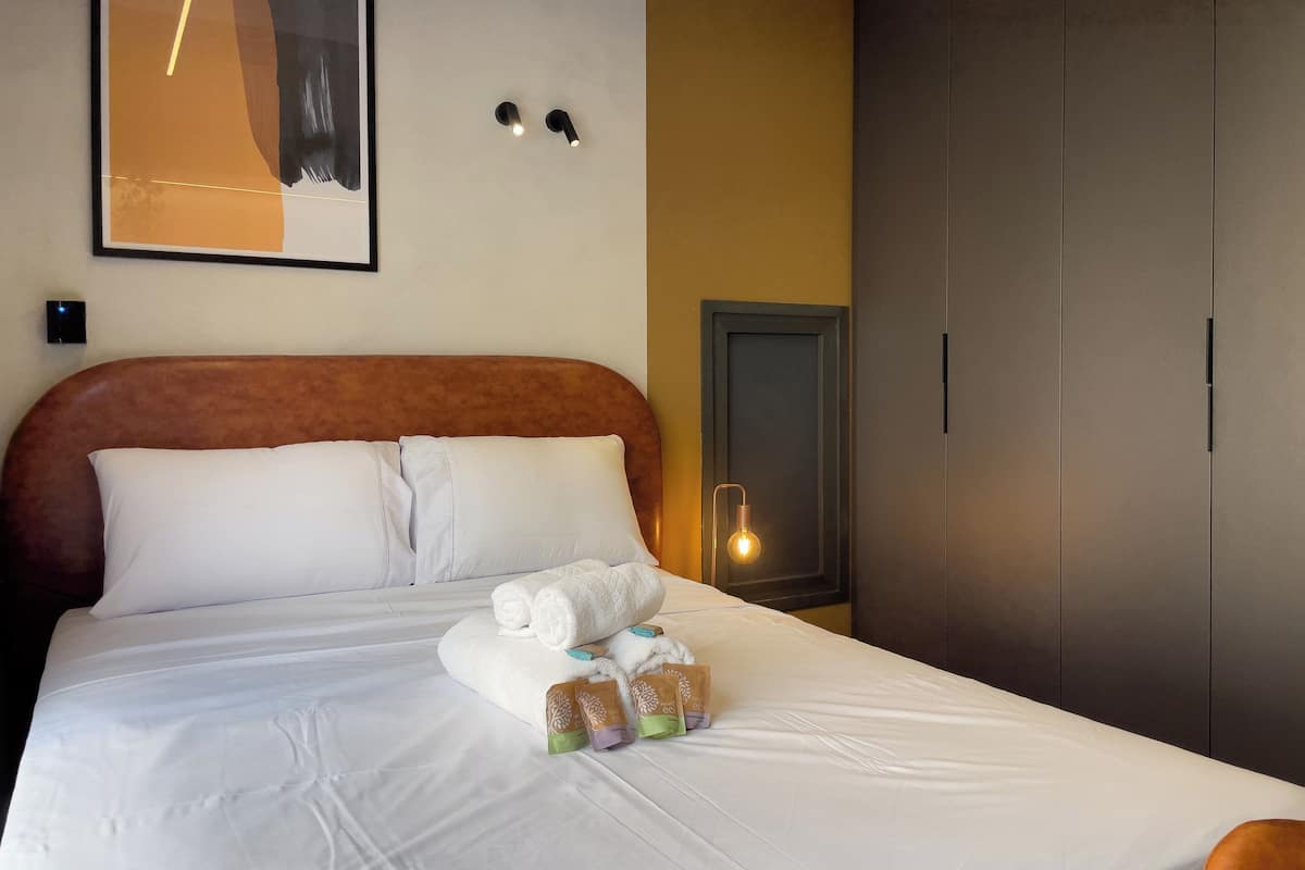 A hotel room with a bed, pillows, a bedside table with a lamp, and a wardrobe.