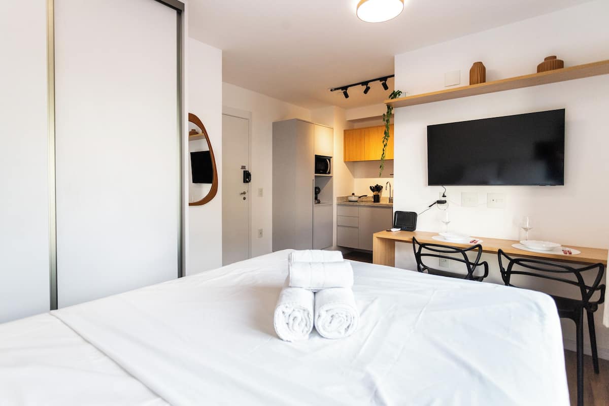 A hotel room with a bed, a desk with a chair, a TV mounted on the wall, and a kitchen area visible through an open door.