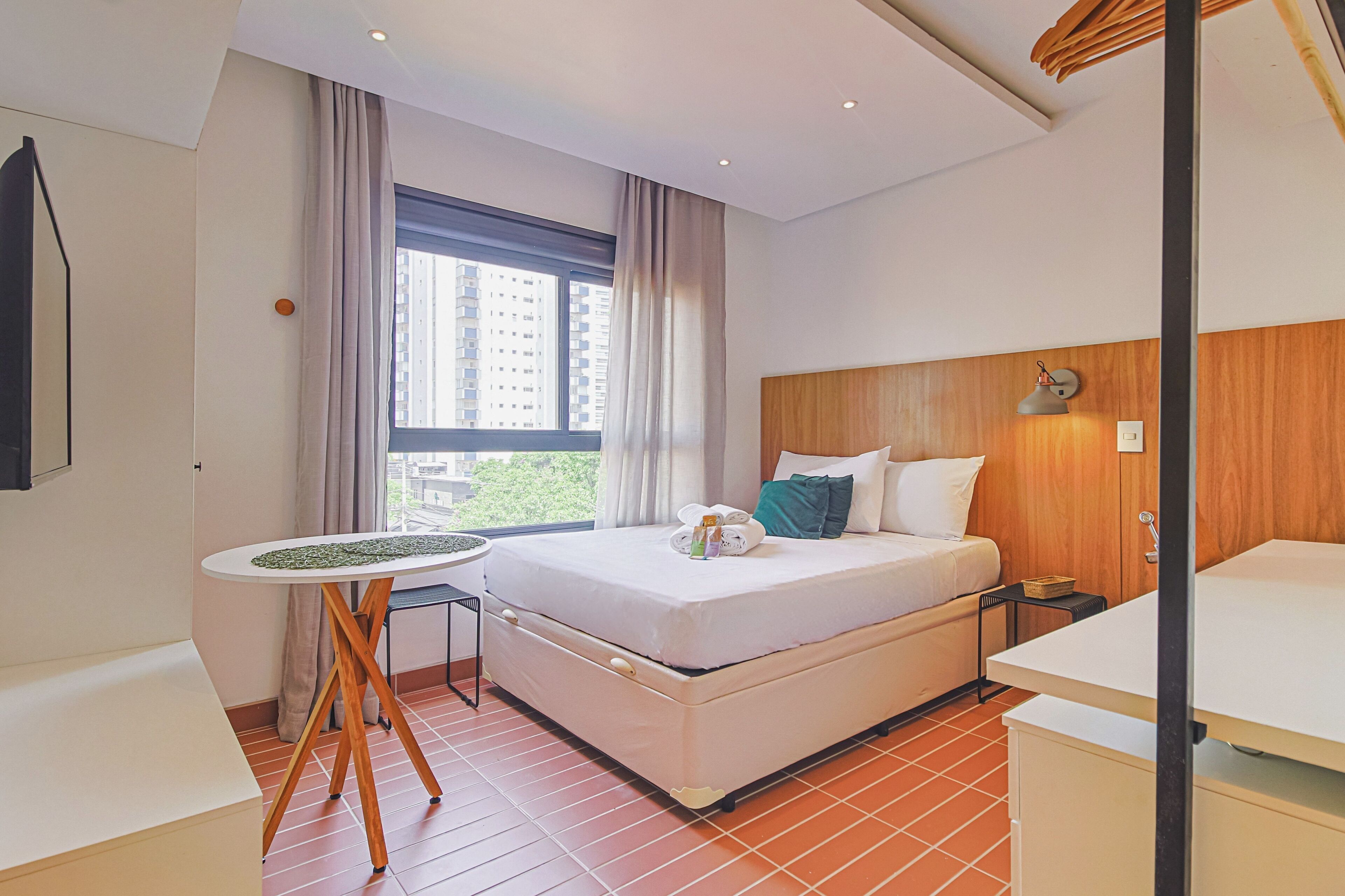 Une chambre d’hôtel avec un lit, une petite table, un bureau, une fenêtre avec des rideaux et une vue sur les bâtiments environnants.