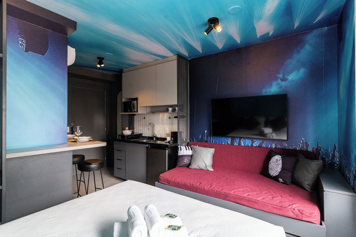 A modern hotel room with a bed, a red sofa, a TV, a kitchen area, and a ceiling with a starry design.