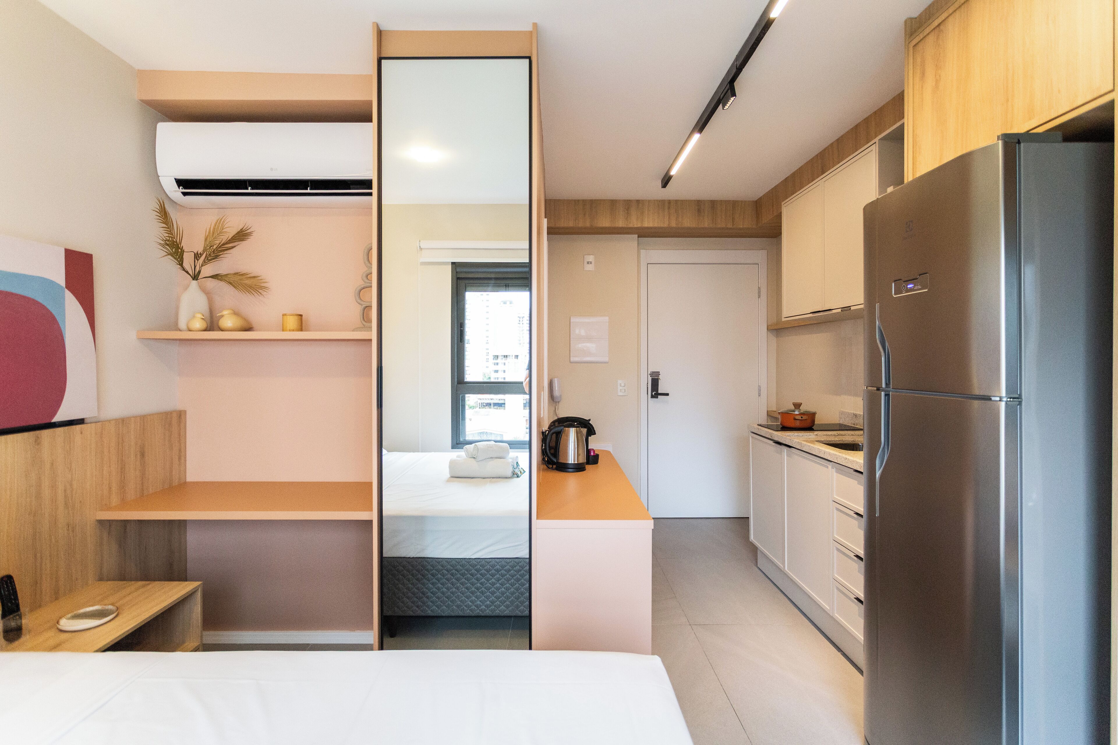 Une chambre d’hôtel moderne équipée d’une kitchenette, d’un lit avec une tête de lit blanche, d’une petite table avec une assiette et d’une plante décorative posée sur une étagère.