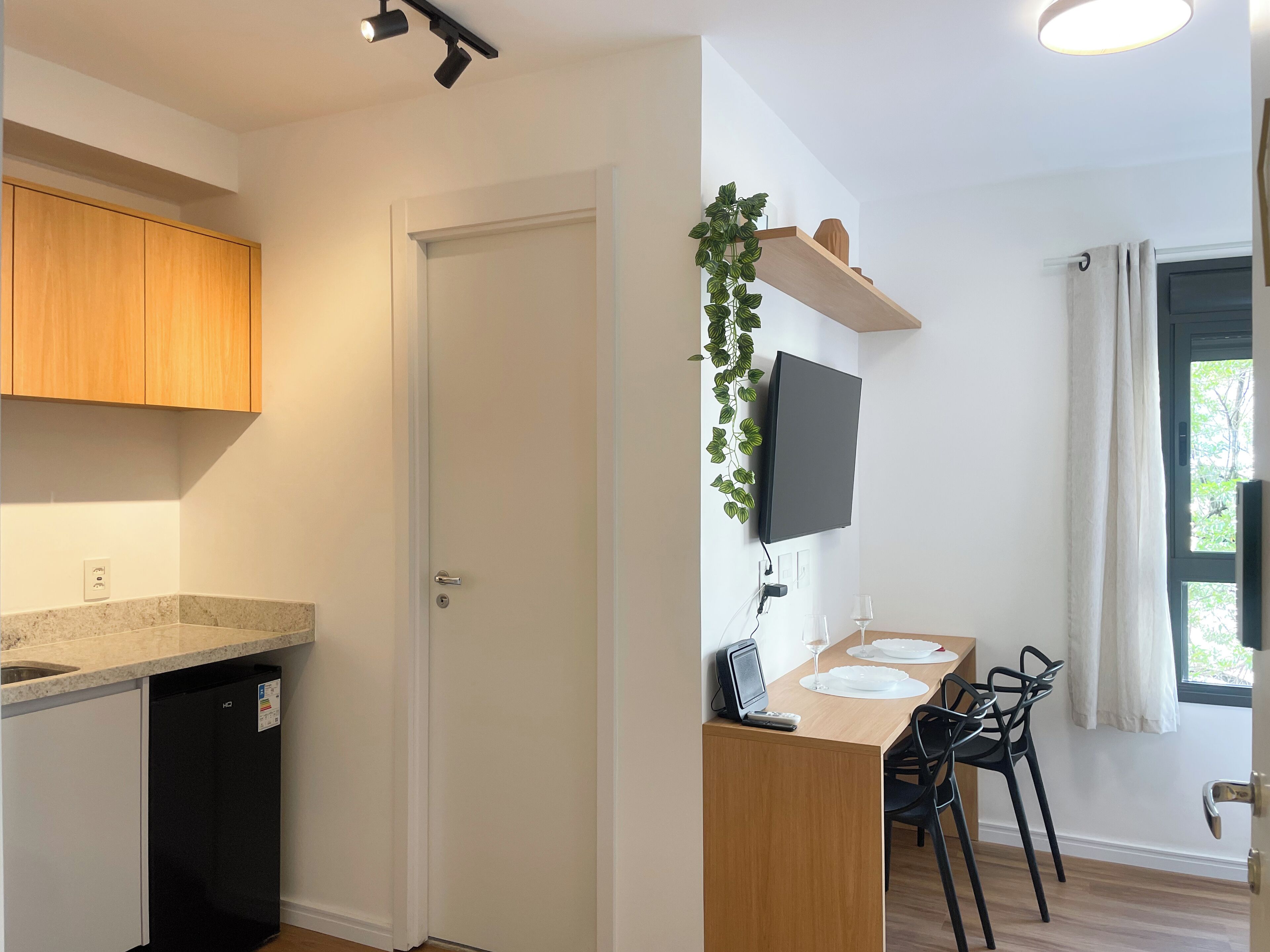 A modern kitchen with wooden cabinets, a built-in dishwasher, a dining area with a table set for two, and a TV mounted on the wall.
