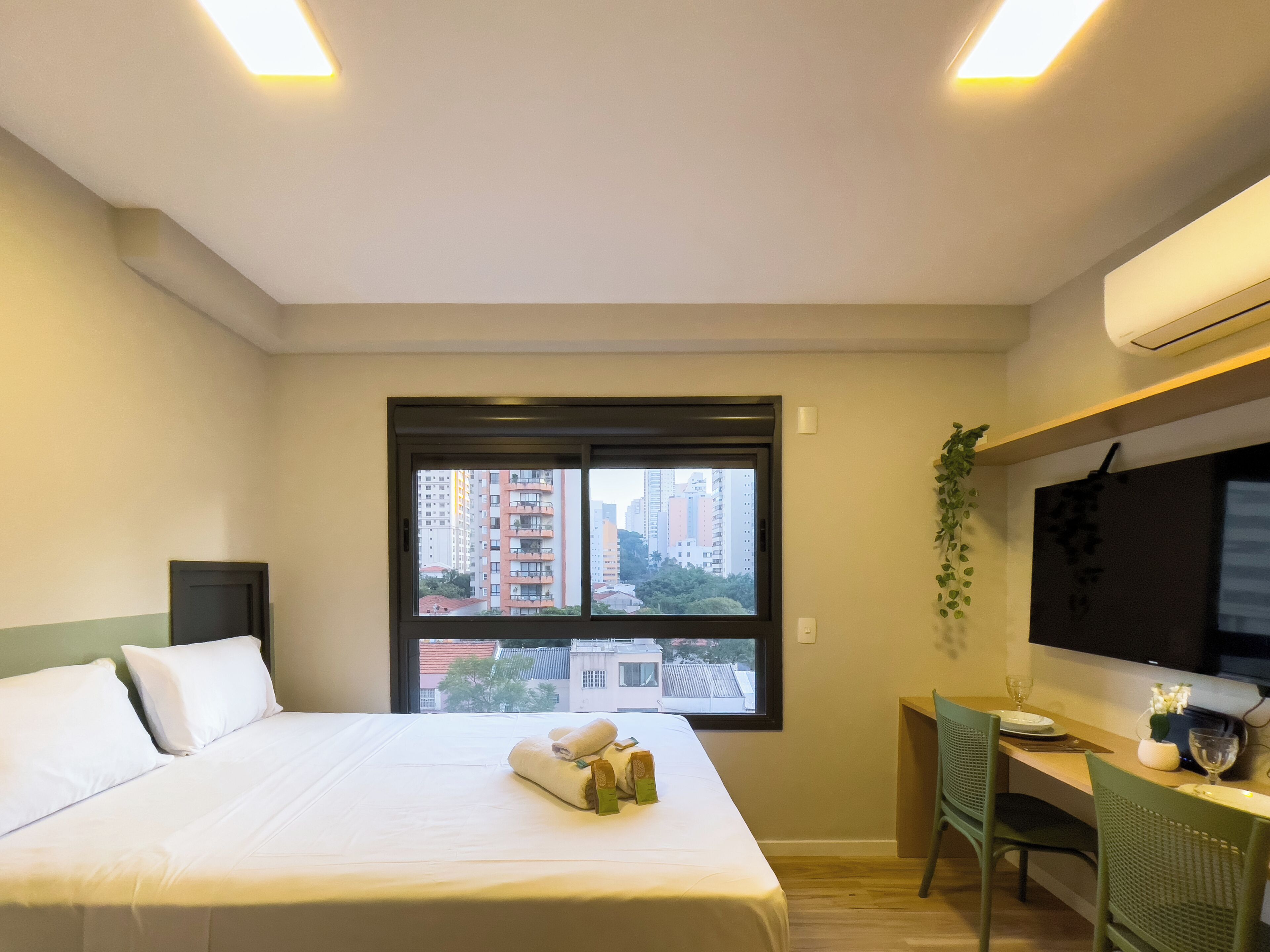Habitació d'hotel amb un llit, un escriptori, una cadira, una finestra amb vista a edificis i un aparell d'aire condicionat.