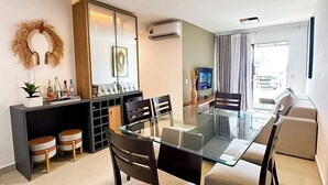 A modern living room with a glass dining table, dark wooden chairs, a black entertainment unit, and a large window with curtains. - Apartment on the sand in the South beaches (Ilheus)