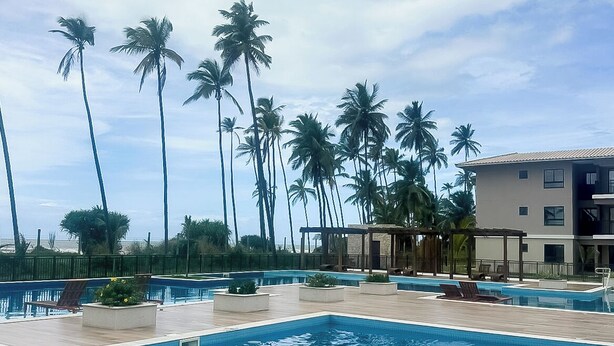 A swimming pool with lounge chairs, palm trees, and a building in the background. - Apartment on the sand in the South beaches (Ilheus)