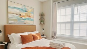 A neatly made bed with white and orange bedding, a bedside table with a lamp, a small shelf with decorative items, and a large window with blinds.