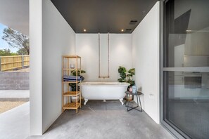 A modern bathroom with a freestanding bathtub, a wooden shelf with towels, and a small table with a potted plant.