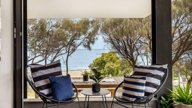A balcony with two chairs and a small table, overlooking a beach with trees and the sea. - Beachfront W/ Breathtaking Views (Blackmans Bay)
