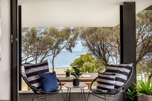 A balcony with two chairs and a small table, overlooking a beach with trees and the sea.