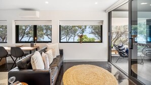 A modern living room with a sofa, chairs, a dining table, and large windows with a view of trees and water. - Beachfront W/ Breathtaking Views (Blackmans Bay)