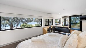 A modern bedroom with a large bed, a desk, and a view of a beach and trees. - Beachfront W/ Breathtaking Views (Blackmans Bay)