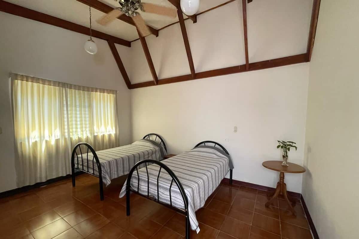 A room with two metal bunk beds, a small wooden table with a plant, a ceiling fan, and a window with curtains.