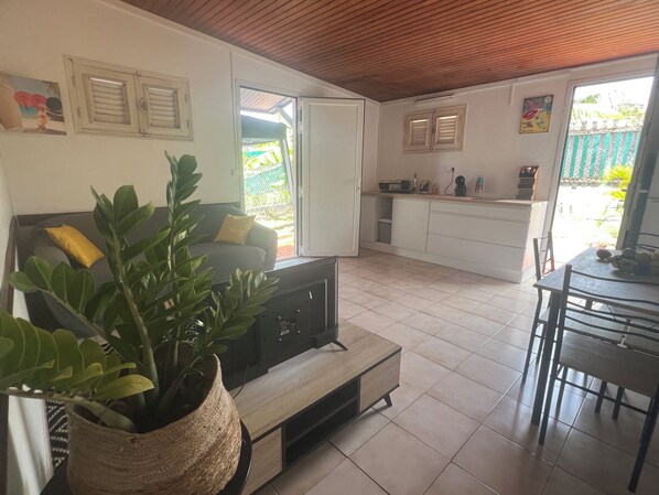 A living room with a sofa, a TV, a dining table with chairs, a kitchen area, and a large potted plant.
