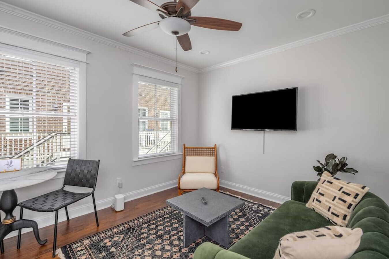 Un salon doté d’un ventilateur de plafond, de deux fenêtres équipées de stores, d’un téléviseur à écran plat, d’un canapé vert, d’un fauteuil noir, d’une petite table basse et d’un tapis à motifs.