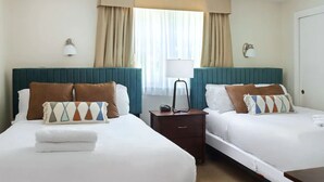 Ein Hotelzimmer mit zwei Betten, jeweils mit einem türkisblauen Kopfteil, weißer Bettwäsche und Zierkissen. Es gibt zwei Nachttische mit Lampen, ein Fenster mit Vorhängen und eine Tür.