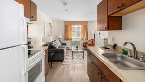 Eine kompakte Küche mit Kühlschrank, Herd und Spüle. Im Wohnbereich befindet sich eine Couch und ein Sessel, sowie ein Fenster mit Vorhängen.