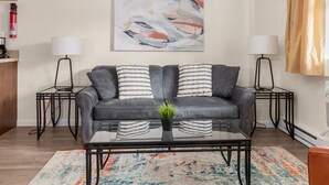 Ein modernes Wohnzimmer mit einem grauen Sofa, zwei Beistelltischen, einer Kaffeemaschine, einem Feuerlöscher und abstrakter Kunst an der Wand.