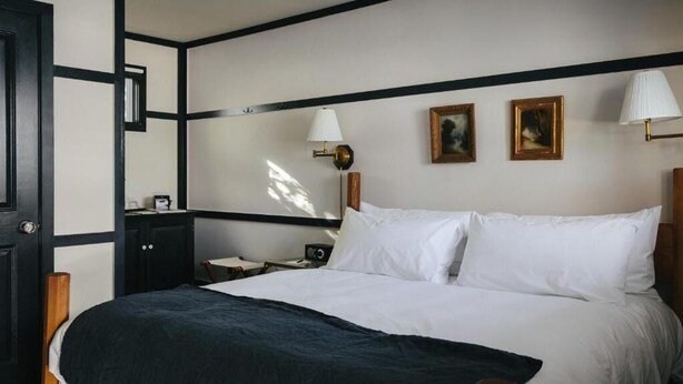 A neatly made bed with white linens and a dark blanket, a bedside table with a lamp, and two framed pictures on the wall.