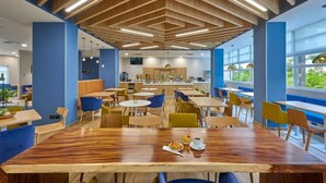 Un comedor moderno con mesas y sillas de madera, una cocina central y grandes ventanales con vistas a zonas verdes.