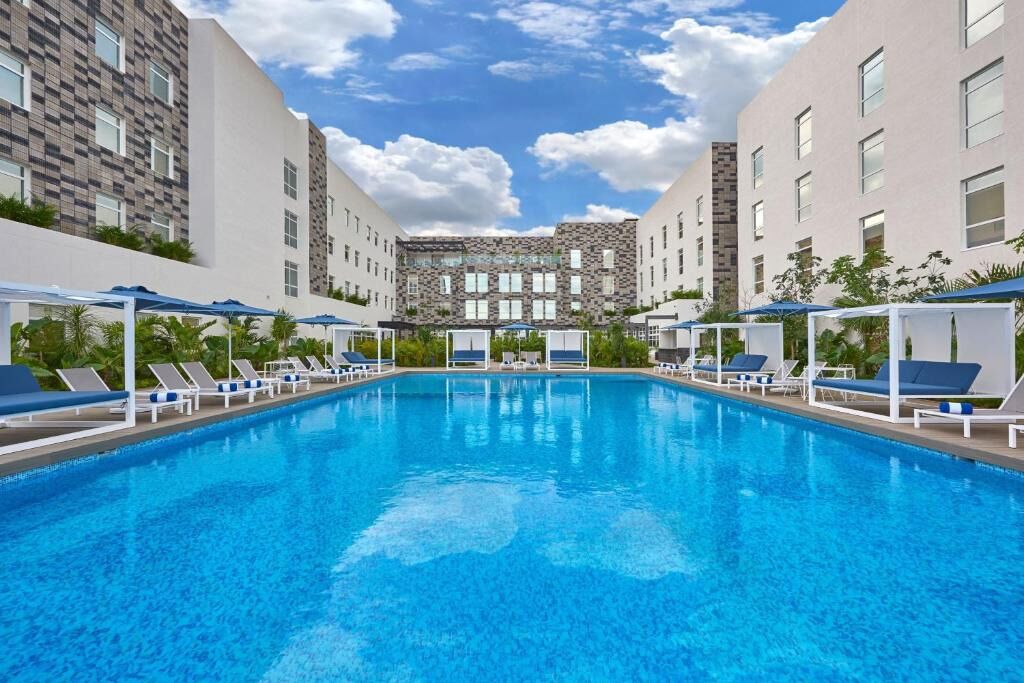 A swimming pool with lounge chairs and umbrellas, surrounded by modern buildings with a checkered facade and a central stone structure.