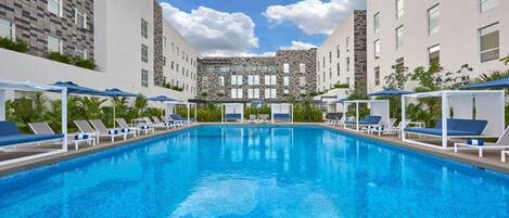 A swimming pool with lounge chairs and umbrellas, surrounded by modern buildings with a checkered facade and a central stone structure.