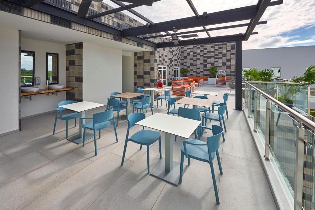 Un espace de restauration extérieur moderne, avec des chaises bleues et des tables blanches, un mur à carreaux et une balustrade en verre donnant sur un paysage urbain.