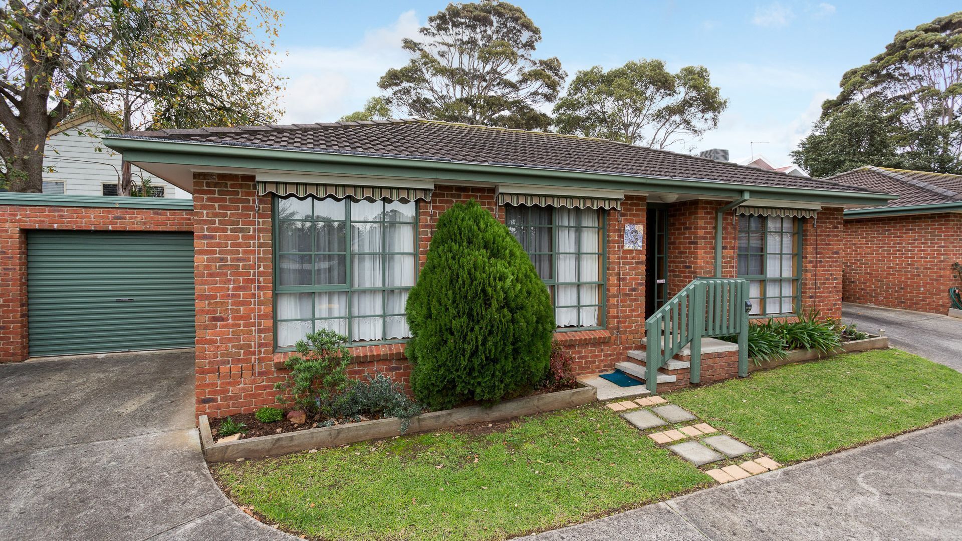 A single-story brick house with a green front door, a small front yard with a trimmed bush, and a driveway leading to a garage.