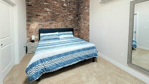 A bedroom with a brick wall, a bed with a patterned bedspread, a nightstand, an air conditioning unit, and a mirror.