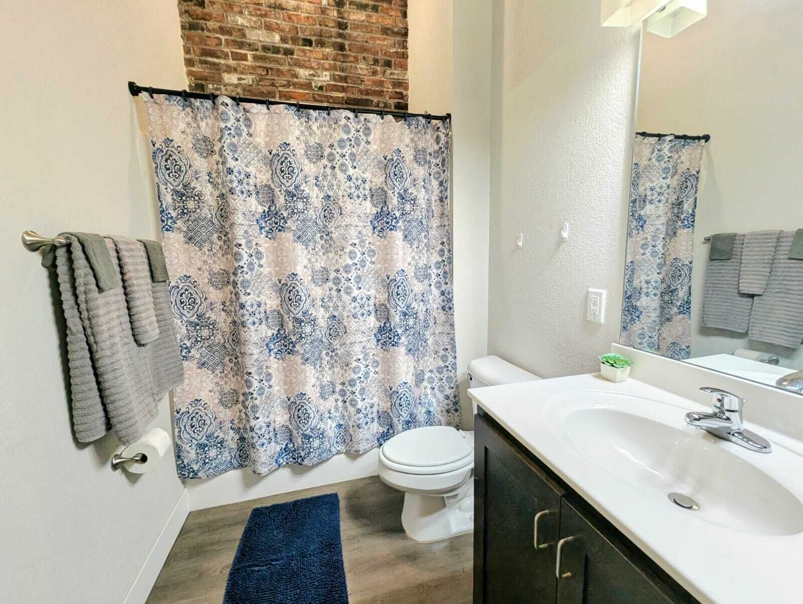 A bathroom with a patterned shower curtain, a white toilet, a sink with a mirror, and a blue rug on the floor.