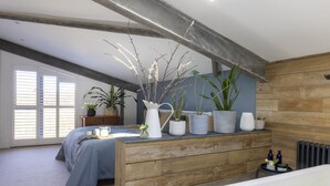 A bedroom with a wooden headboard, a bed with a blue bedspread, a bedside table with a white vase and plants, and a window with blinds.