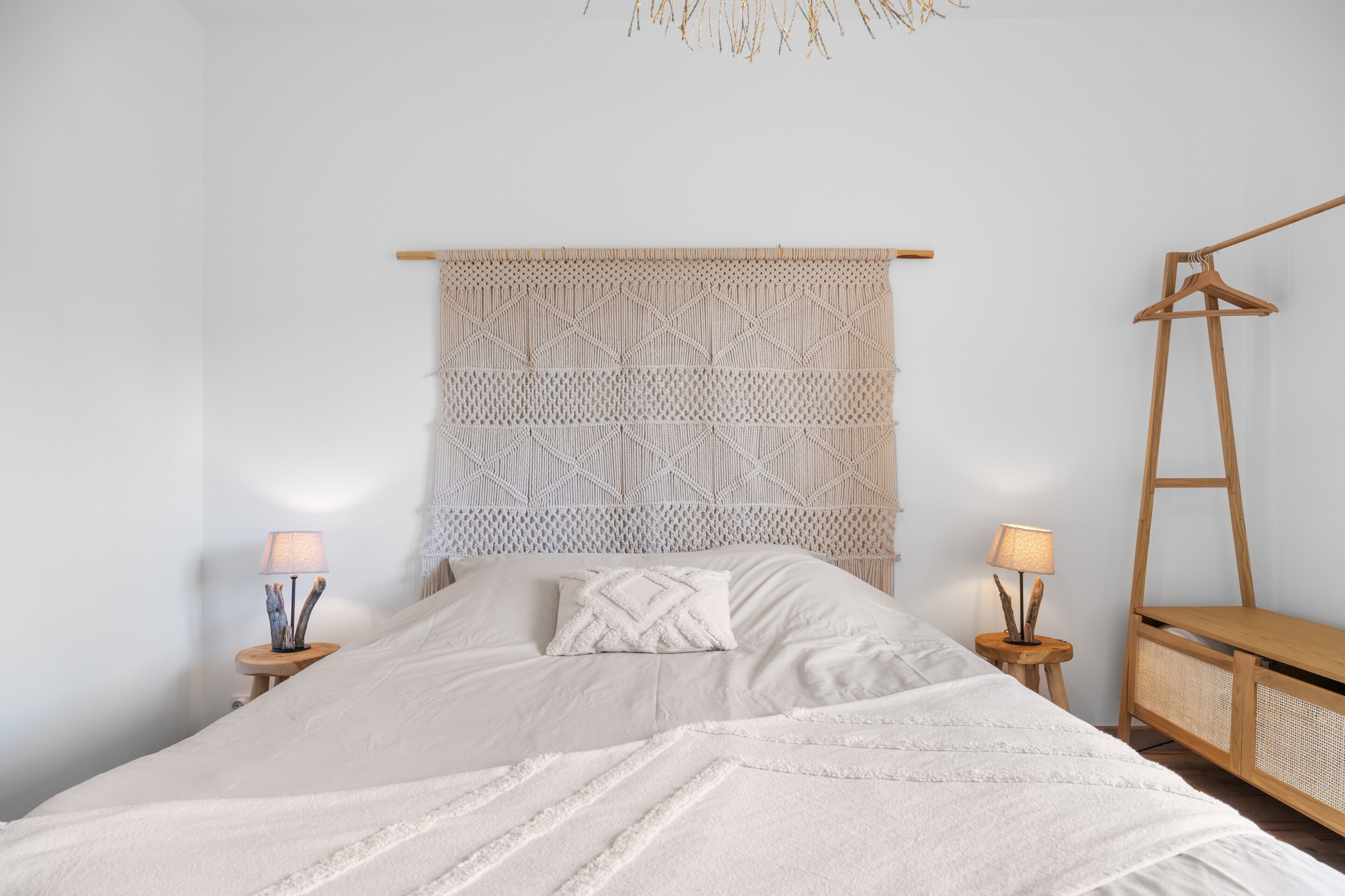 Un dormitorio ordenado con cama, dos mesitas de noche con lámparas, una escalera de madera y un cabecero tejido.