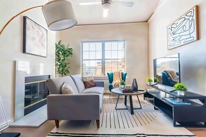 A modern living room with a sofa, coffee table, and TV stand, featuring a large window, artwork, and a plant. - Boulevard Stays Spacious 1BR King Suite Pool, Gym Access & Free Parking (Irving)