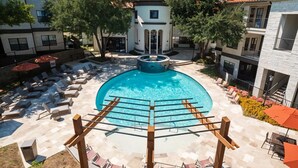 A swimming pool with sun loungers and a pergola in the foreground, surrounded by buildings with balconies and a small tower structure. - Boulevard Stays Spacious 1BR King Suite Pool, Gym Access & Free Parking (Irving)