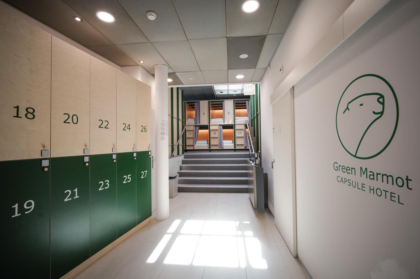 A hotel corridor with lockers numbered 18-27, a staircase leading to rooms, and a wall with the Green Marmot Capsule Hotel logo.