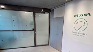 A modern hotel lobby with a glass door entrance, a welcome sign, and a wall-mounted logo for Green Marmot Capsule Hotel.
