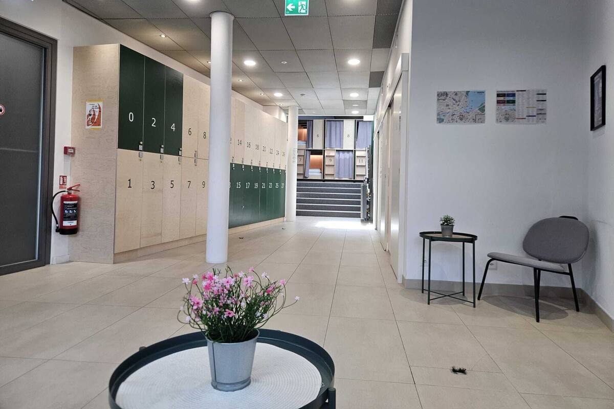 A hotel lobby with lockers, a fire extinguisher, a plant, a chair, and a staircase leading to another floor.