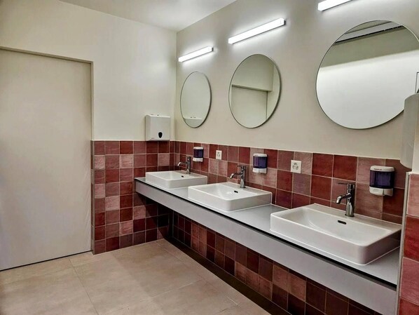 Un baño moderno con tres lavamanos, cada uno con un espejo encima. Hay dispensadores de jabón en la pared y una puerta a la izquierda.