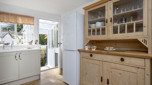 A kitchen with white cabinets, a wooden cabinet with glass doors, a white refrigerator, and a window with a view of houses and greenery. - Withycombe is a beautifully presented dog-friendly terraced cottage for six guests (Seaton)