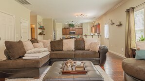 A living room with a sectional sofa, a coffee table with decorative items, and a kitchen in the background. - Serene Family Hideaway 4Br w Pool Table (Fortson)