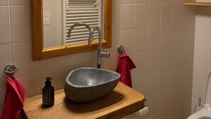 Een badkamer met een spiegel met houten lijst, een stenen wastafel, een kraan, een rode handdoek, een zeepdispenser en een toilet.