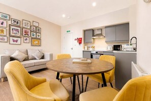 A modern living room with a round wooden table, yellow chairs, a grey sofa, and a kitchen area with grey cabinets and a white countertop.